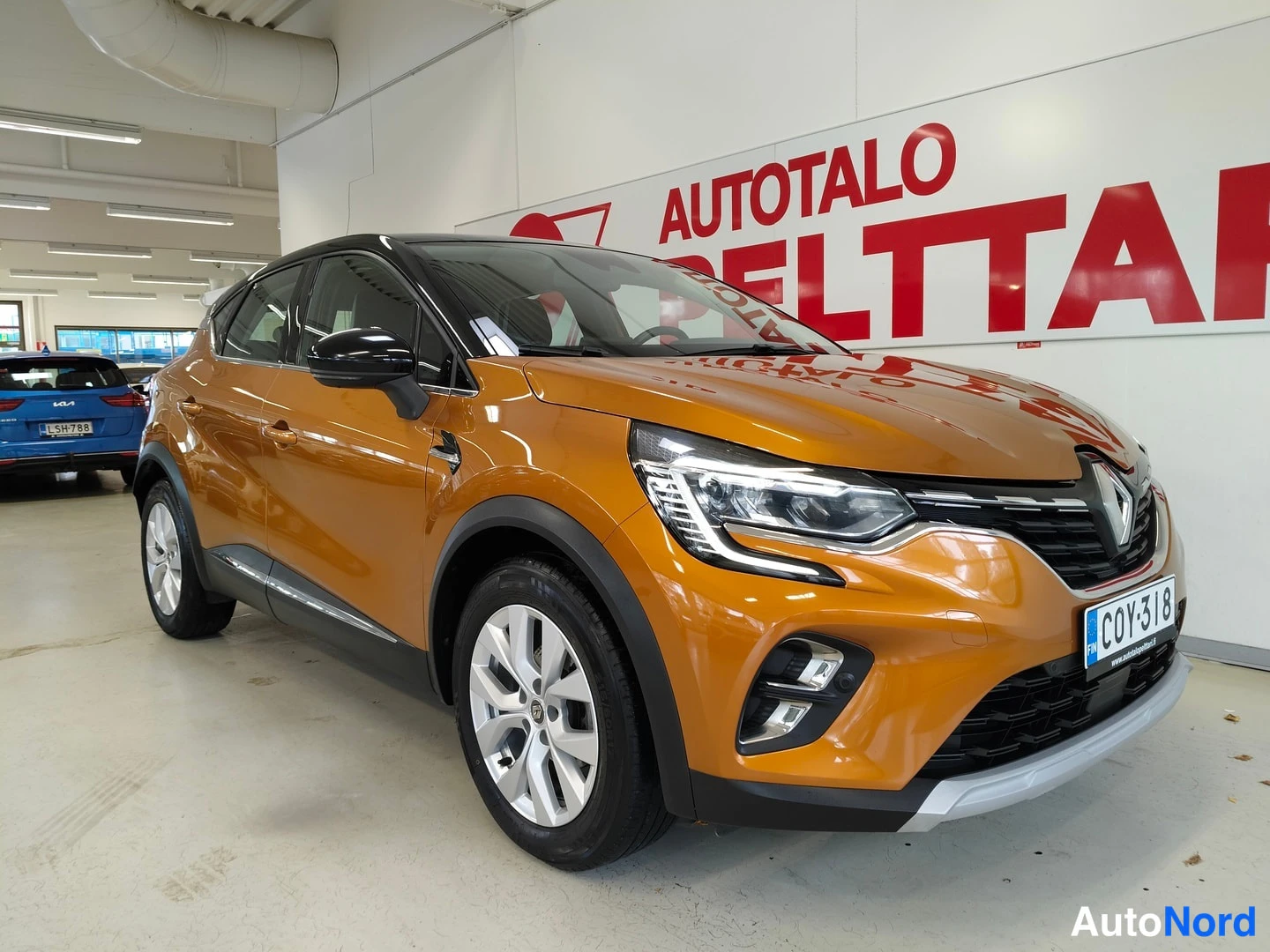 Etuviisto, nokka oikealle: Oranssi Renault Captur etuviistosta kuvattuna sisätiloissa; rekisterikilpi ja Renault-merkki etusäleikössä näkyvät.