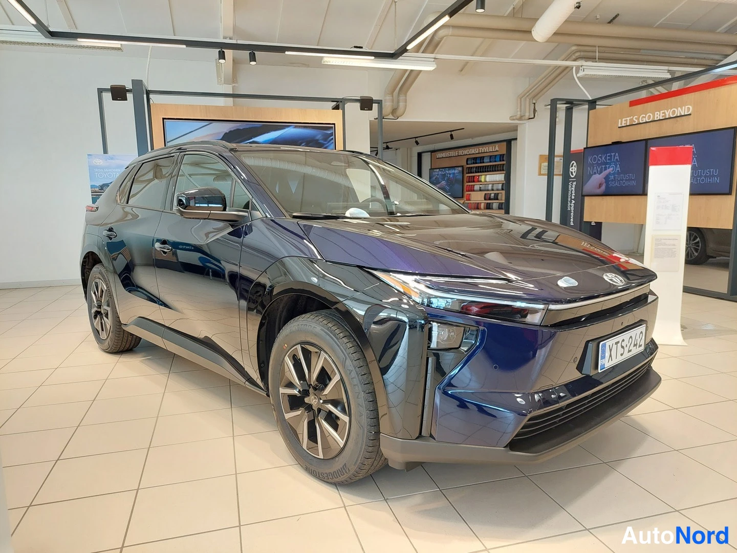 Etuviisto, nokka oikealle: Toyota BZ4X tummansinisenä, keula oikealla vasemmalle kääntyneenä auton ollessa autosalon näyttelytilassa; rekisteritunnus.