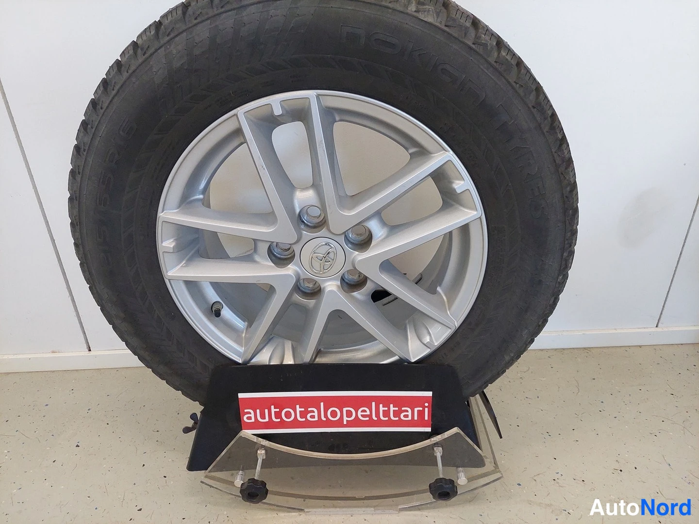 Yksityiskohta: Irrotettu Toyota-kehäpyörä hopeavanteella, kokkoja Nokian Talvi-Hakkapeliitta 8 -renkaassa, esillä 'autotalopeltari'-tekstikyltillä.