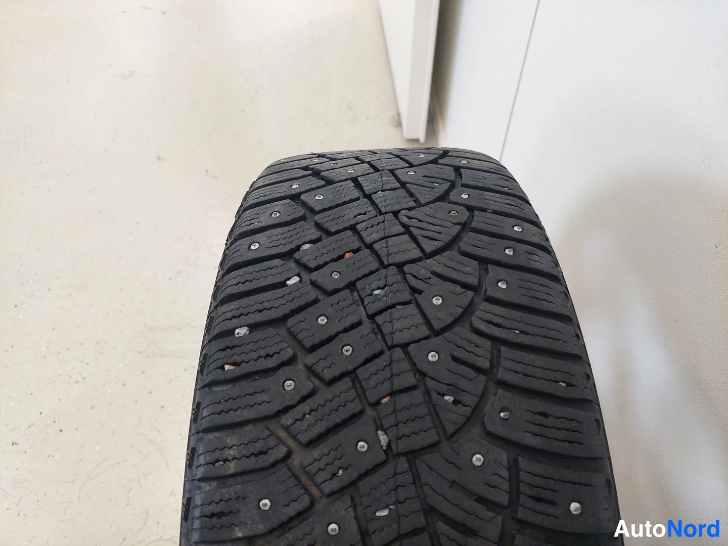 Yksityiskohta: Kenkkäkuvioinen nastarenkaan kulmapinta näkyy lähikuvassa harmaalla lattialla.