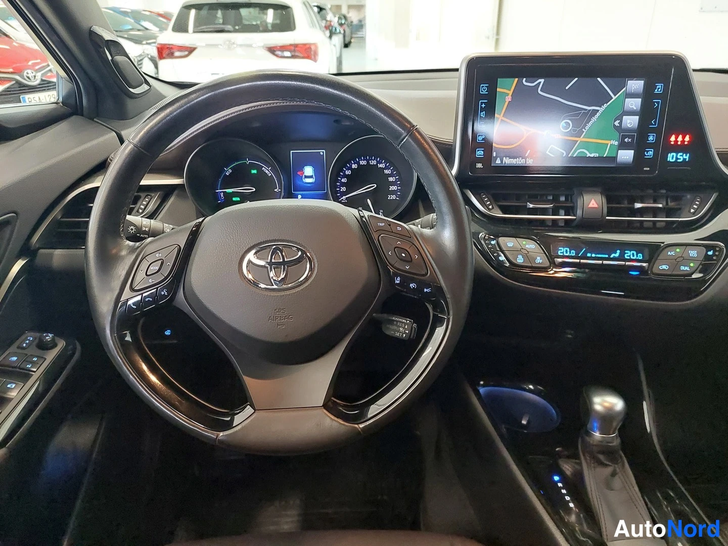Muu: Ohjauspyörän lähikuva: Toyota-merkki, mittaristo (hybridimittari ja nopeusmittari), infotainment-näyttö kartalla ja ilmastointipaneeli, arvo 20,0 molemmin puoli