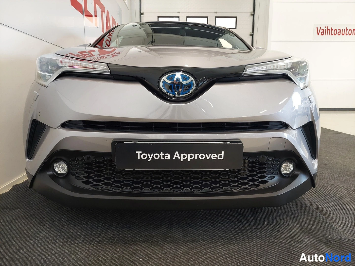 Edestä: Auton keula suoraan edestä, sininen Toyota-hybridimerkki, Toyota Approved -kyltti etupuskurissa, hopeanharmaa koripi.