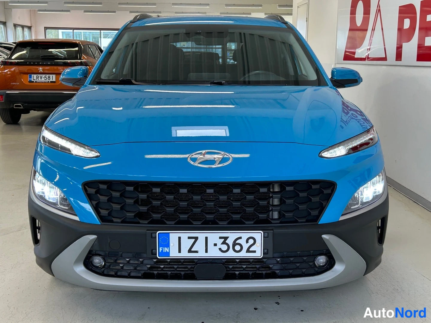 Edestä: Sinisenä autossa Hyundai Kona edestä päin, rekisterikilpi, ajoneuvosali sisätiloissa.