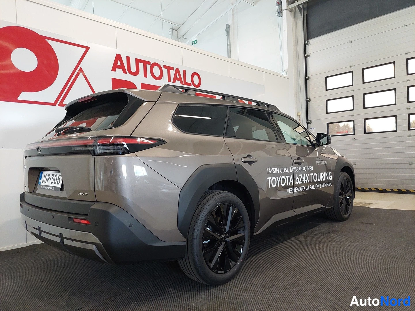 Takaviisto: Auton takakulmanäkymä: ruskeakarmaa korivaatteessa oleva Toyota bZ4X Touring, jonka takapuskuri ja mustat kevytmetallivanteet näkyvät sisätiloissa.
