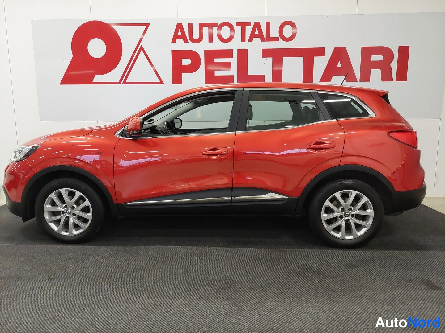 Sivulta: Punaisen Renault Kadjarin kylkikuva sisätiloissa, valkoinen seinä taustalla.