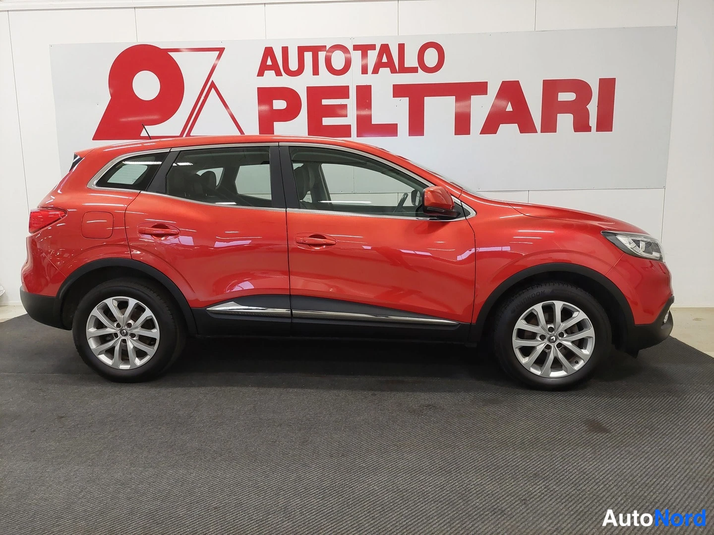 Sivulta: Punaisen Renault Kadjarin sivuprofiili sisätiloissa, Autotalo Pelttari -taustakyltti takana.