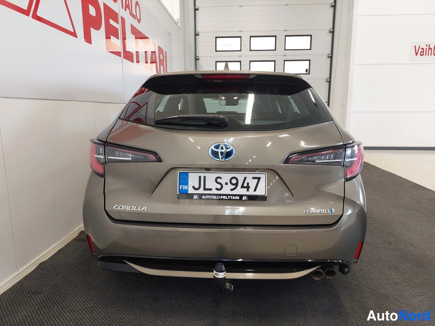Takaa: Ruskeanharmaan Corolla-station wagonin takaosa, rekisterikilpi, HYBRID-merkintä ja Corolla-teksti takaluukissa, vetokoulukytkin taakse jääneessä puskurissa.