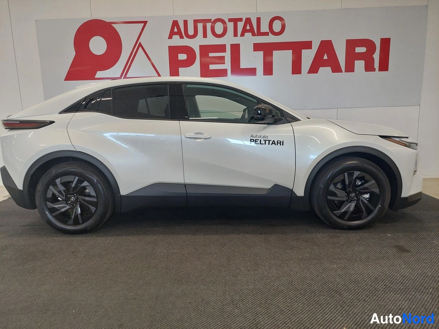 Etuviisto, nokka oikealle: Valkoinen Toyota C-HR sivulta oikeasta kyljestä, mustat kevytmetallivanteet, Autotalo Pelttari -teksti ovessa.