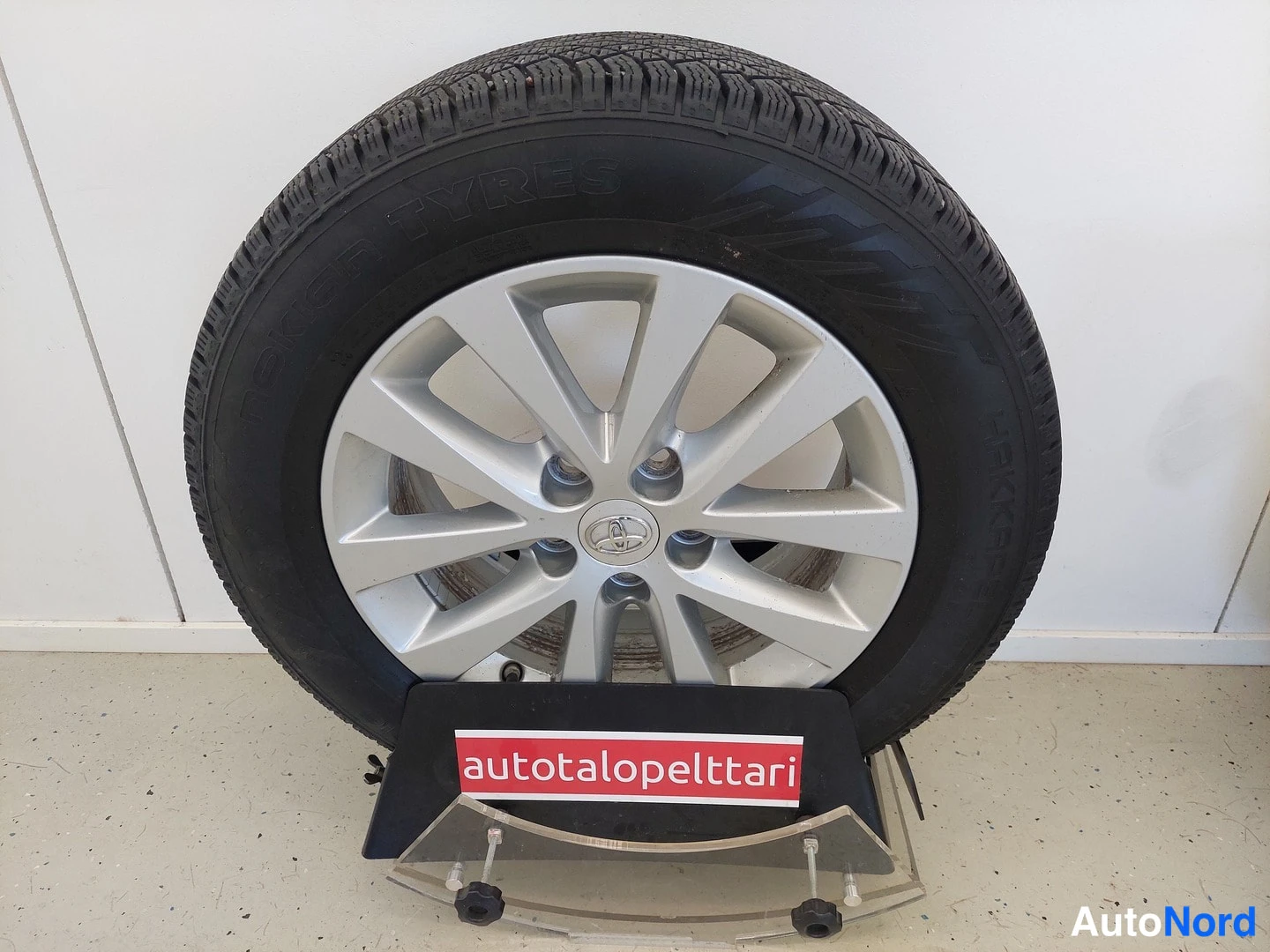Muu: Toyota-merkki varustettu hopeavanne ja Nokian kesärengas seistään, taustalla autotalopeltari-kyltti.