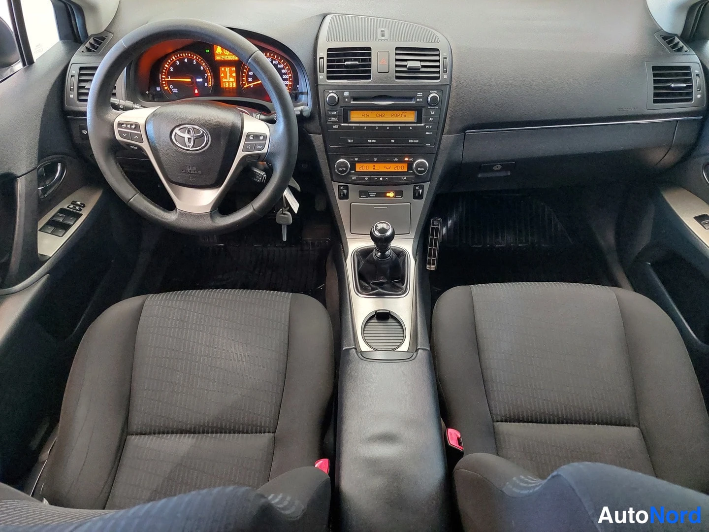 Muu: Toyota Avensisin matkustamo ylhäältä kuvattuna: kaksi eteenpenkkiä, manuaalivaihteisto, Toyota-ohjauspyörä ja kojelauta.