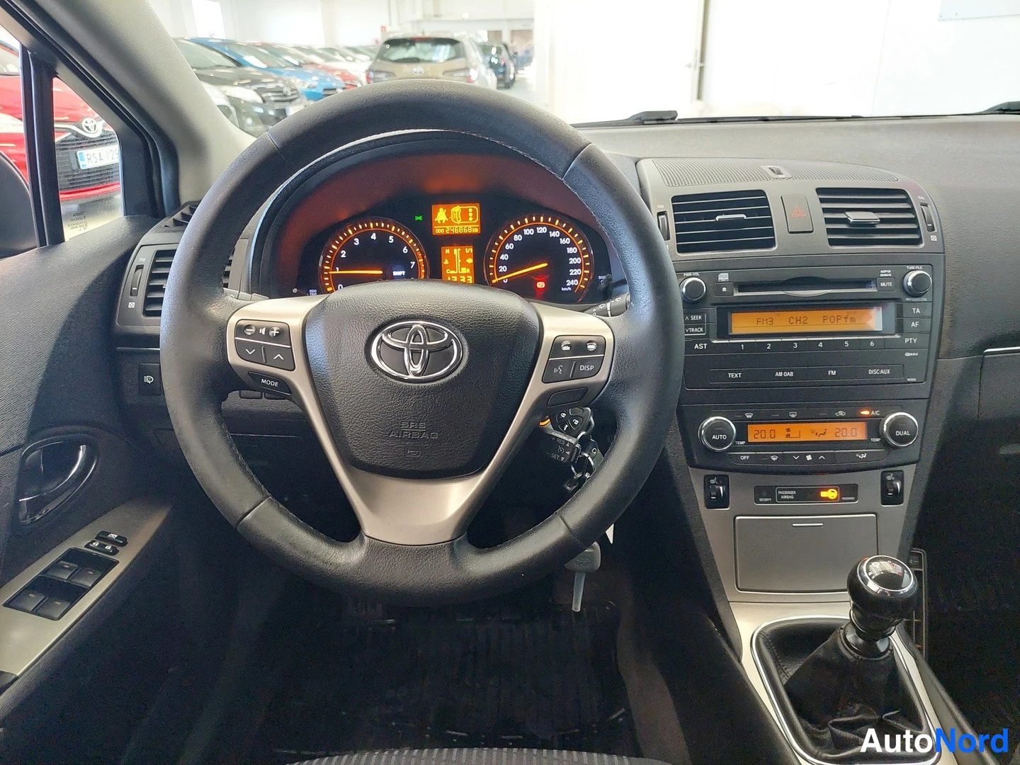 Muu: Toyota Avensisin kuljettajan näkymä: Toyota-ohjauspyörä, mittaristo ja radiopaneeli, vaihteenkeksi näkyvissä.