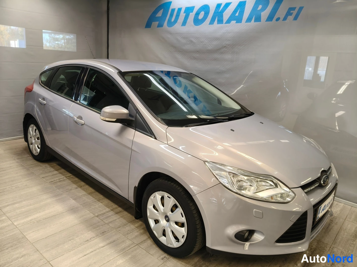 Etuviisto, nokka oikealle: Hopeanharmaa Ford Focus -henkilöauto autosalin lattialla, etukolmannelsku: keula ja ajovalot oikealla, auton kylki ja takapyörä vasemmalla.