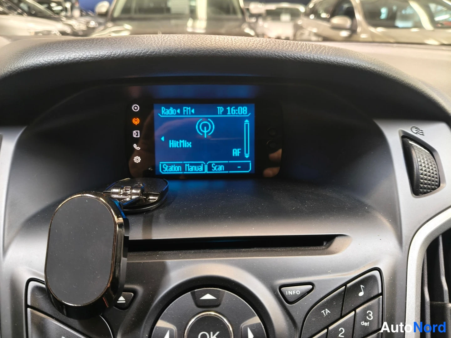 Yksityiskohta: Lähikuva keskityksennäytöstä, johon näkyy radioasema FM1, signaalin voimakkuus ja kello (16:08). Kuvaan heijastuu valoja ja takaa näkyvissä autoja.