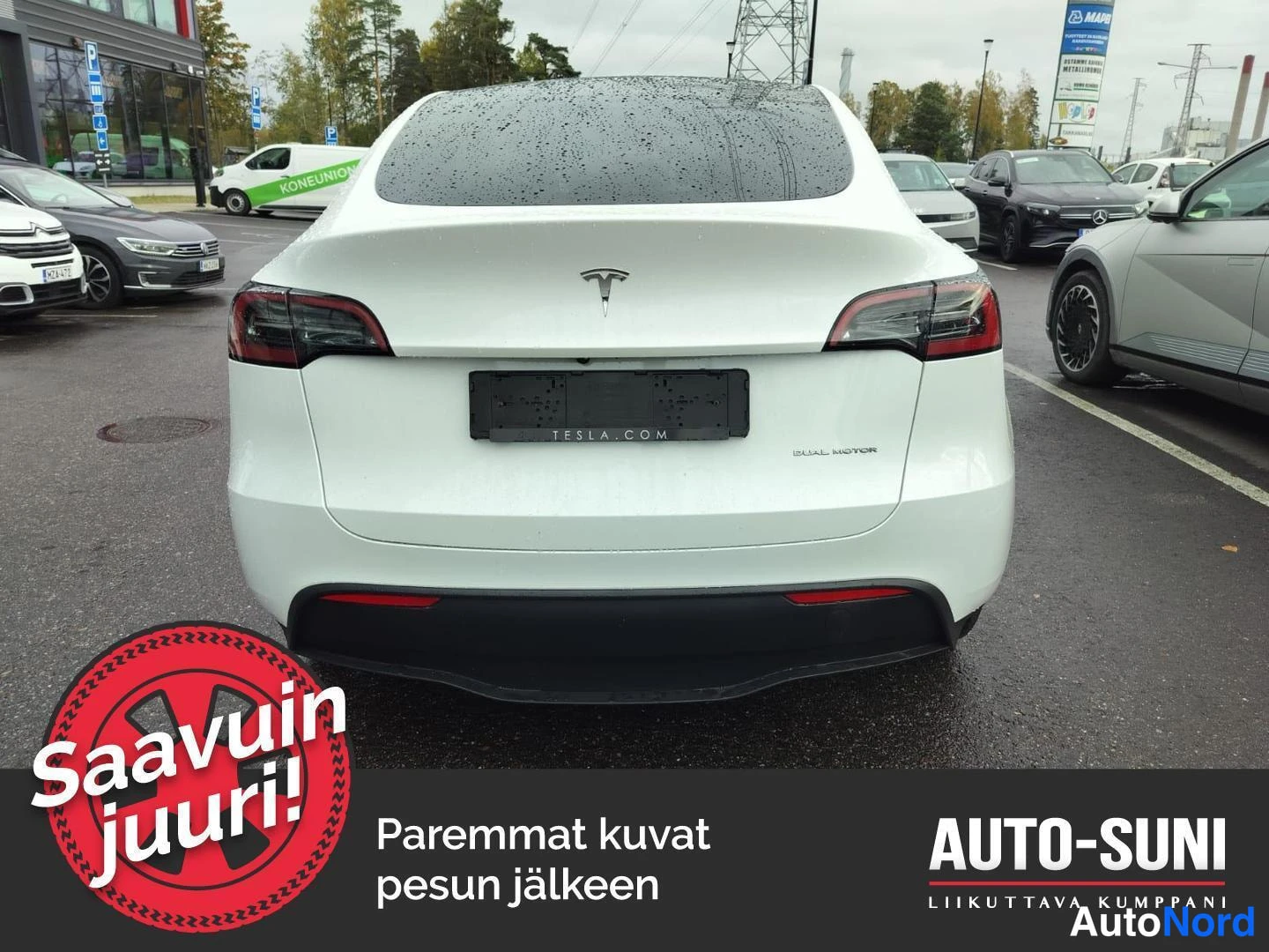 Takaa: Teslan takanäkymä: valkoinen crossover, punaiset takavalot, TESA-teksti rekisterikilven alla, märkä parkkipaikka myyntialueella.