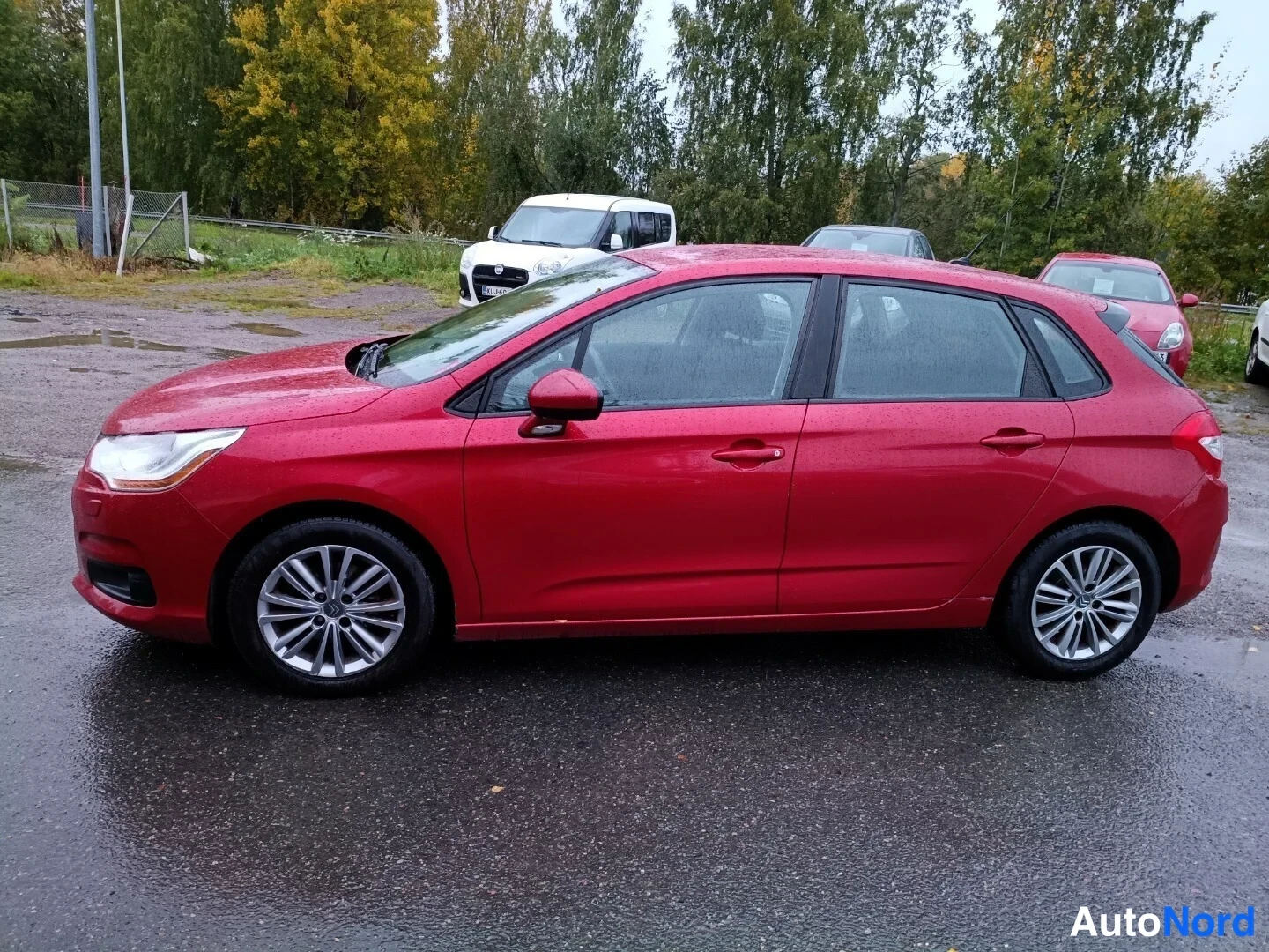 Sivulta: Punainen viisiovinen viistoperäinen auto sivulta, sadevesipisarat korissa, märkä katu ja puusto taustalla.