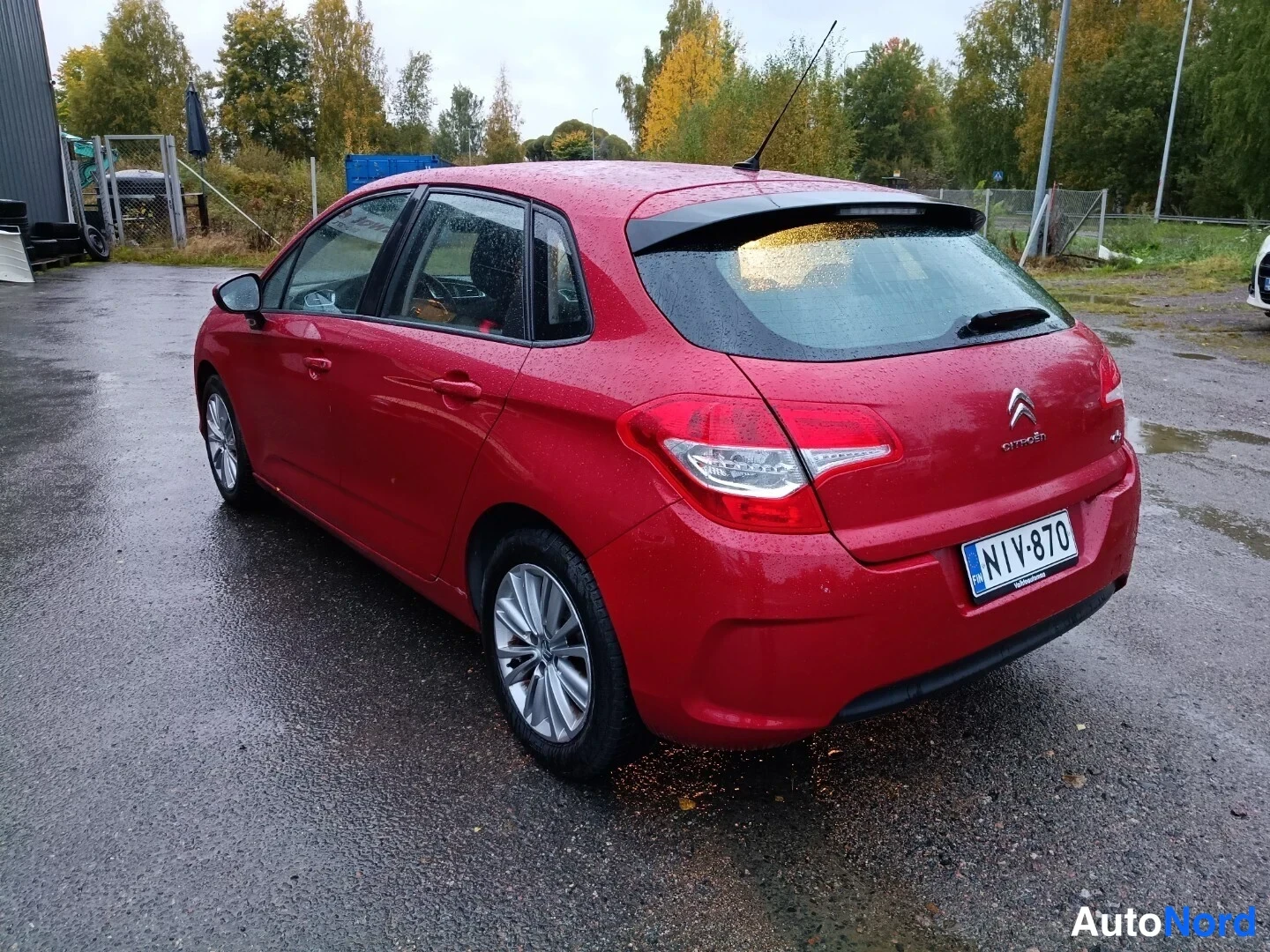 Takaviisto: Punaisen auton takakulma, Citroën-merkki takaluukussa, rekisterikilpi, taustalla halleja ja kenttä.