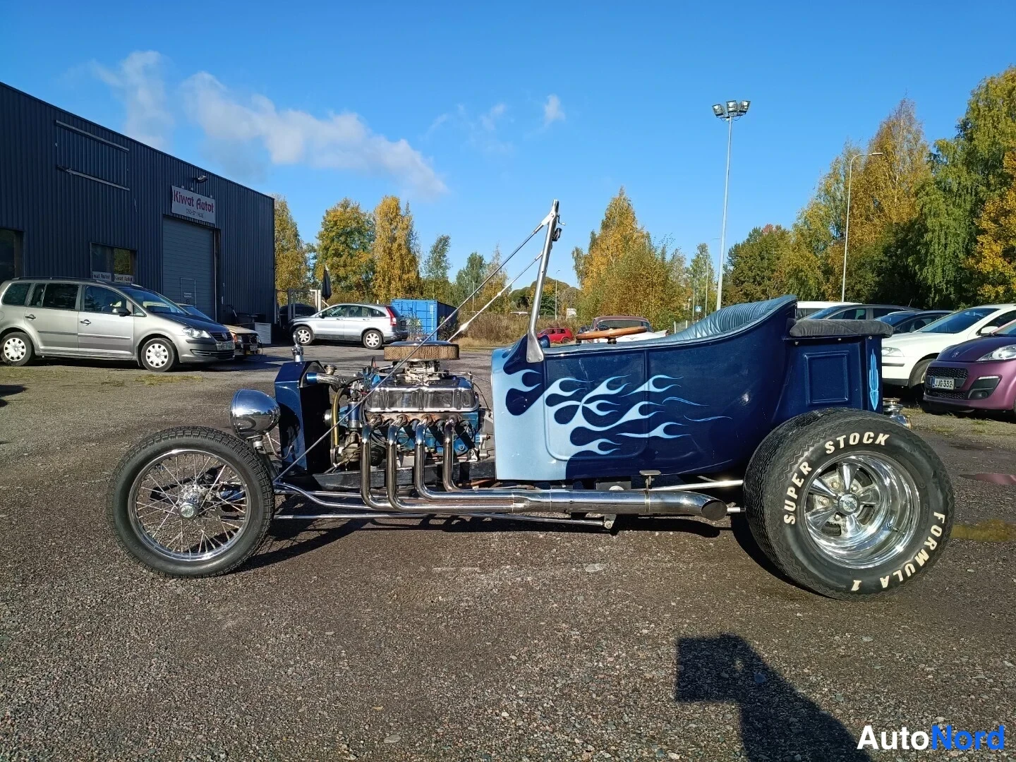 Sivulta: Ford T-bucket sivulta nähtynä, tummansininen kori vaaleansinisellä liekkimaalauksella, paljas V8-moottori ja pakosarjet näkyvissä.