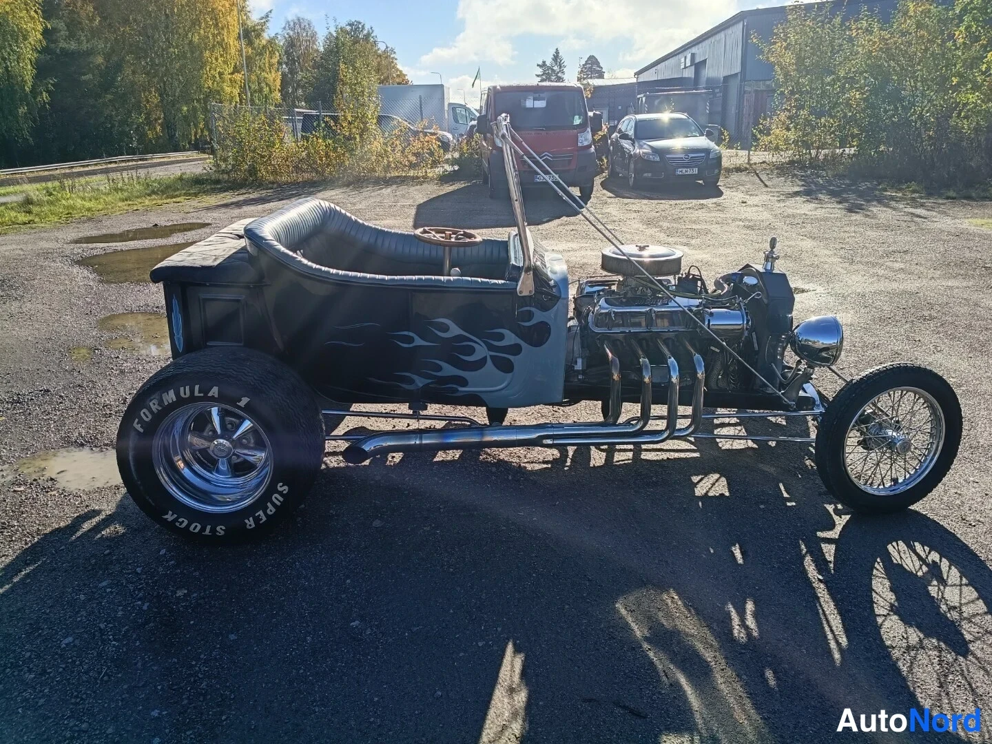 Etuviisto, nokka oikealle: Musta avoin hot rod, jonka sivuprofiili ja keula ovat kuvassa; V8-moottori ja kroomatut pakkoputket näkyvät avoimessa konepellissä.