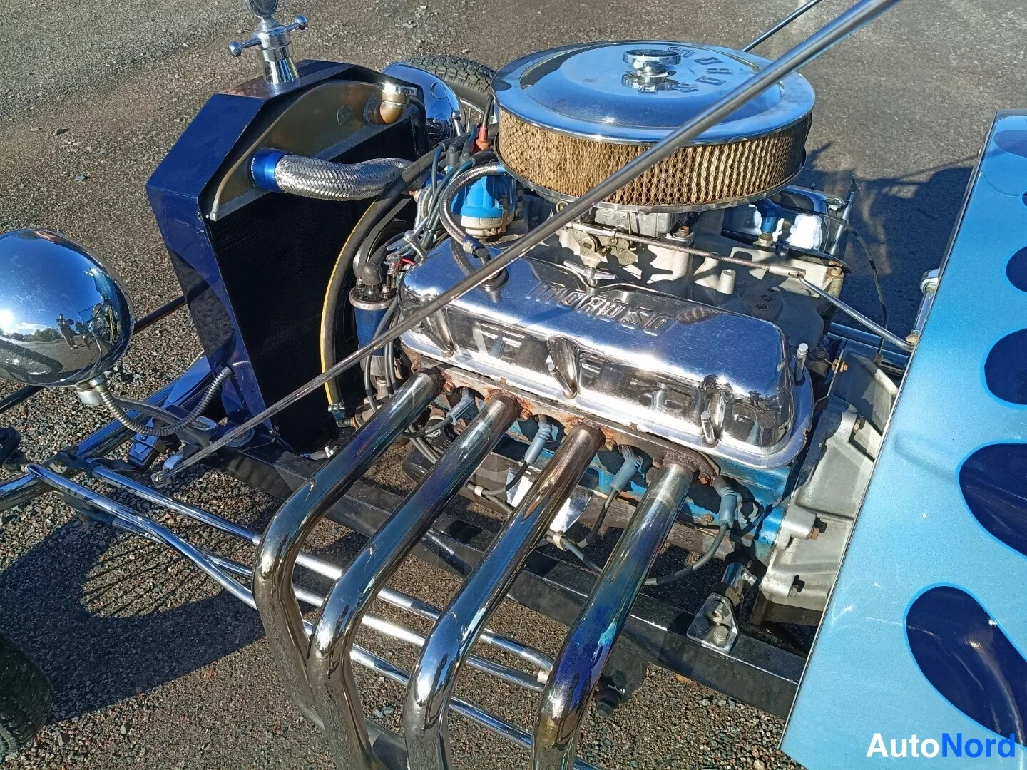 Muu: Lähikuva avoimen hot rod -autoon asennetusta moottorista, jossa kiiltävä kromiset imu- ja pakosarvet, ilmansuodatin ja sininen moottorilohko; vasemmalla kromine