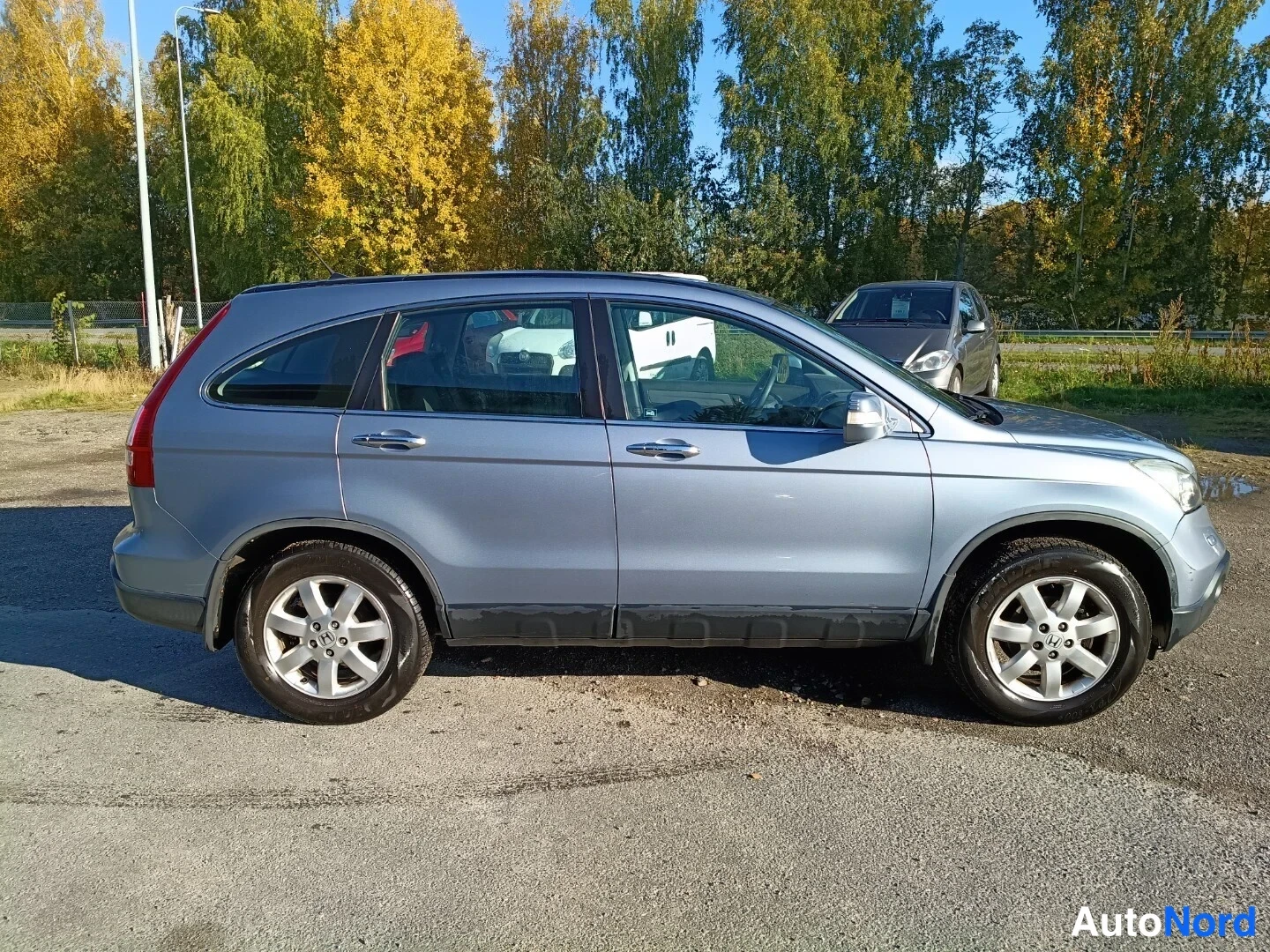 Sivulta: Honda CR-V:n sivukuva, auton kylki ja molemmat pyörät näkyvät hiekkaisella pihalla puiden edessä.
