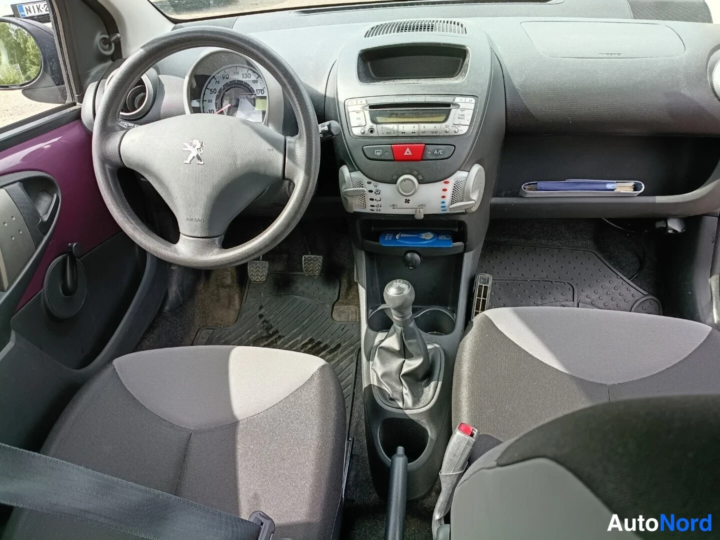 Edestä: Peugeot 107:n ohjaamo etupäältä: ratissa Peugeot-logo, keskikäyttötaulu radion ja A/C-painikkeella, manuaalivaihtehanla ja kaksi etuistuinta.