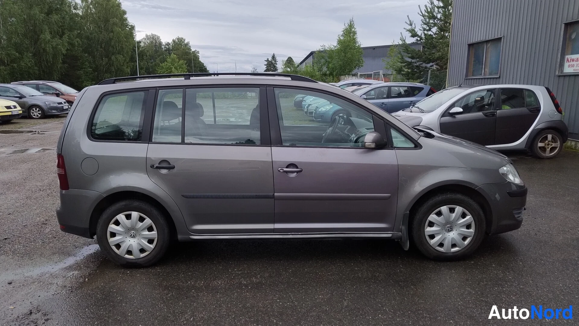 Etuviisto, nokka oikealle: Volkswagen Touranin vasen kylki kolmannelta neljäsosakulmalta; harmaa koripohja, kattokaiteet ja lievä lika ovipaneeleissa märällä parkkipaikalla.