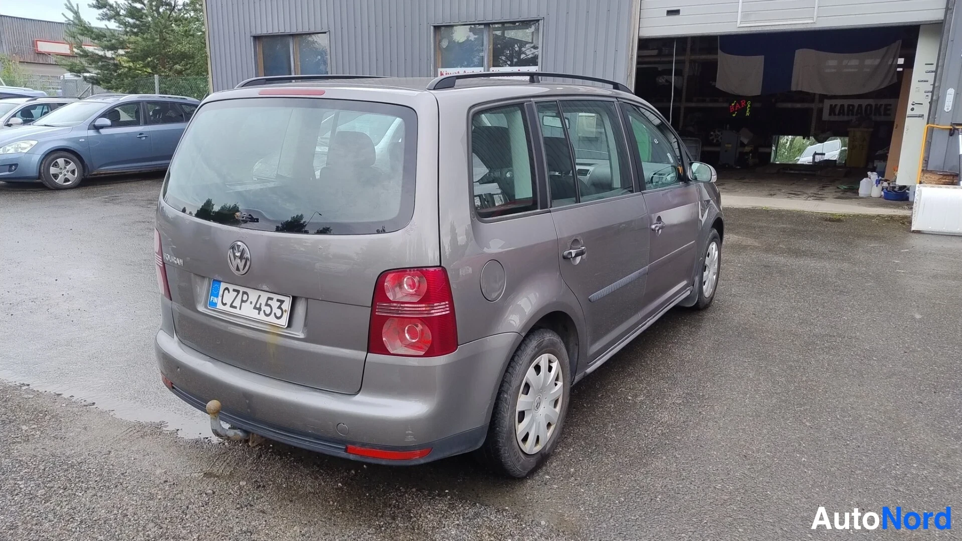 Takaviisto: Volkswagen Touranin takaosa kolmannelta neljäsosakulmalta; harmaa takaosa, rekisterikilpi ja vetoaisa takapuskurissa autotallin edustalla.