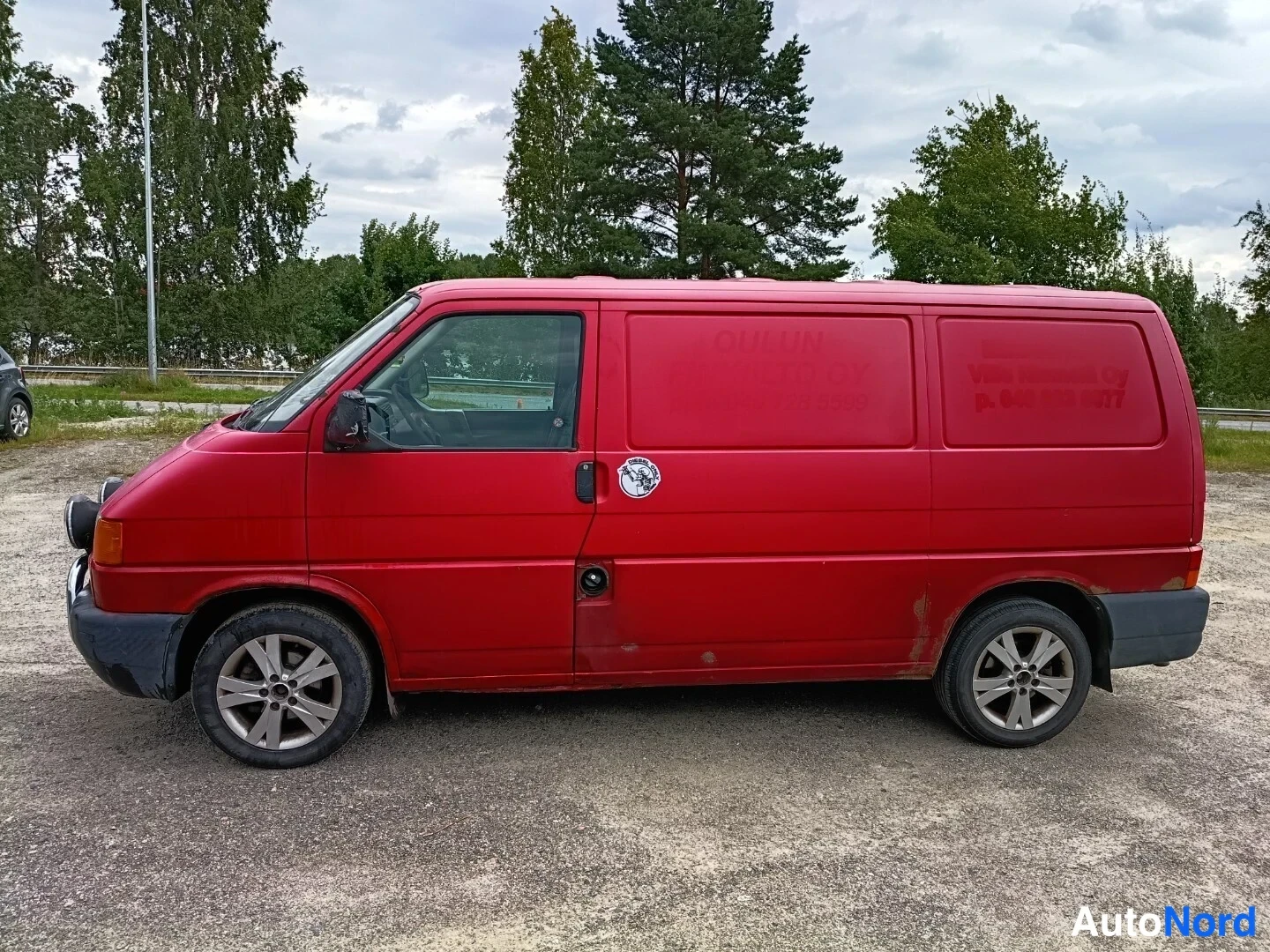 Sivulta: Punaisen Volkswagen Transporter -pakettiauton sivuprofiili vasemmalta, kirjoitusteksti ovessa haalistunut.