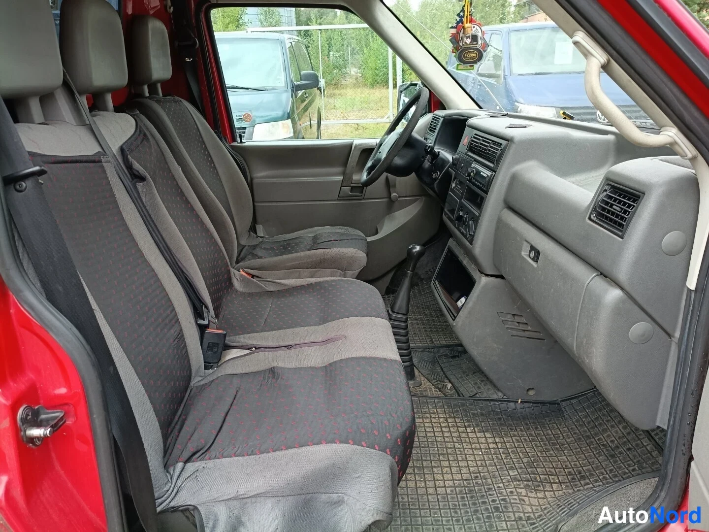 Muu: Volkswagen Transporterin etuistuimet ja kojelauta ovelta kuvattuna; kangasistuimet, turvavyöt ja vaihteenvalitsin näkyvät.