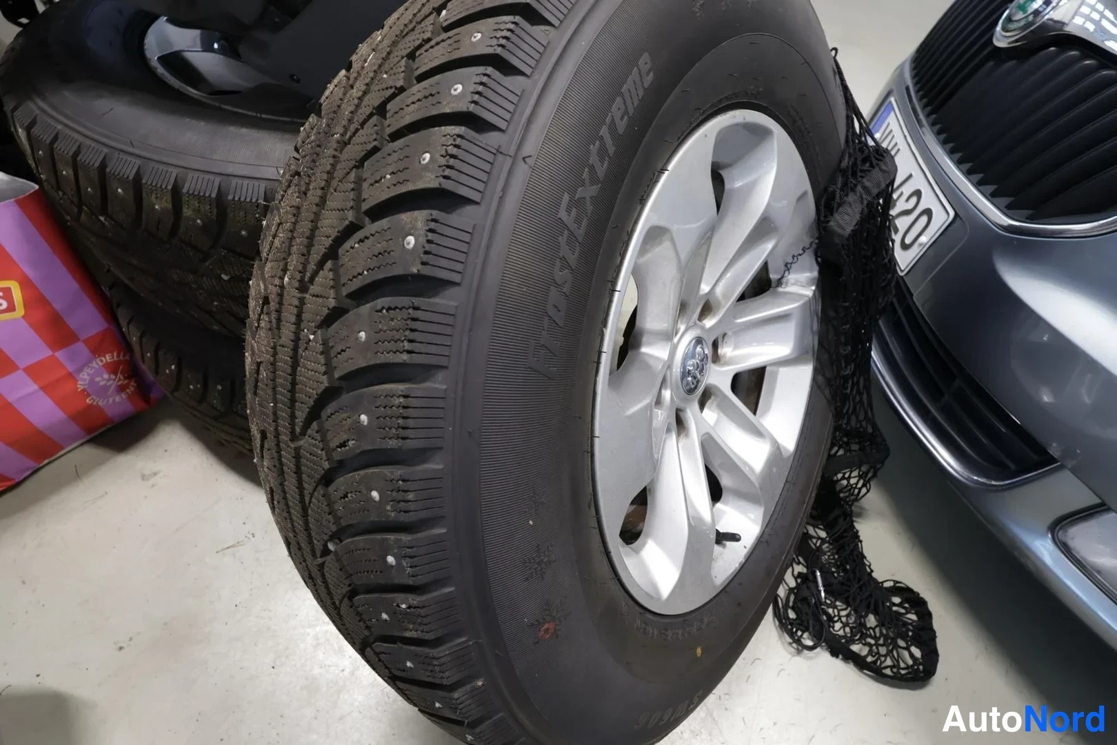 Yksityiskohta: Hopeavanne hiottu alumiinivannes ja naulattu talvirengas Nokian, vieressä harmaa henkilöauto.