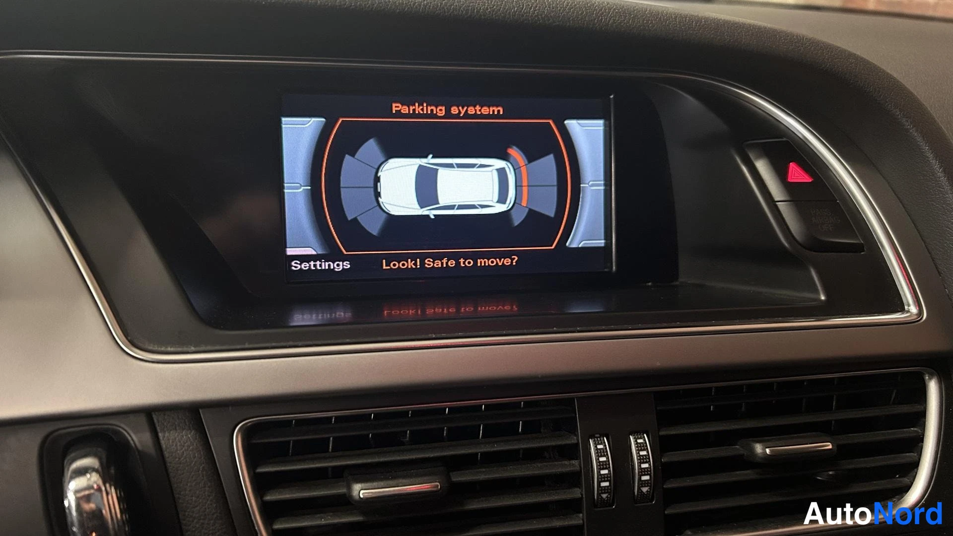 Muu: Kojelautaan upotetun näytön kuva, jossa näkyy pysäköintijärjestelmän grafiikka auton ympärillä ja teksti 'Parking system', 'Settings' ja 'Look! Safe to move?'.