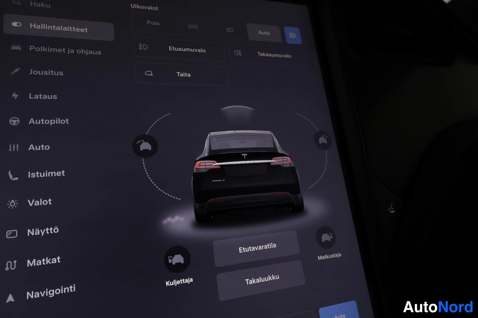 Yksityiskohta: Auton näytön kuvakaappaus: ulkovalojen asetukset ja avattavien osien valikko, Taakse kuvattu musta Tesla Model X.