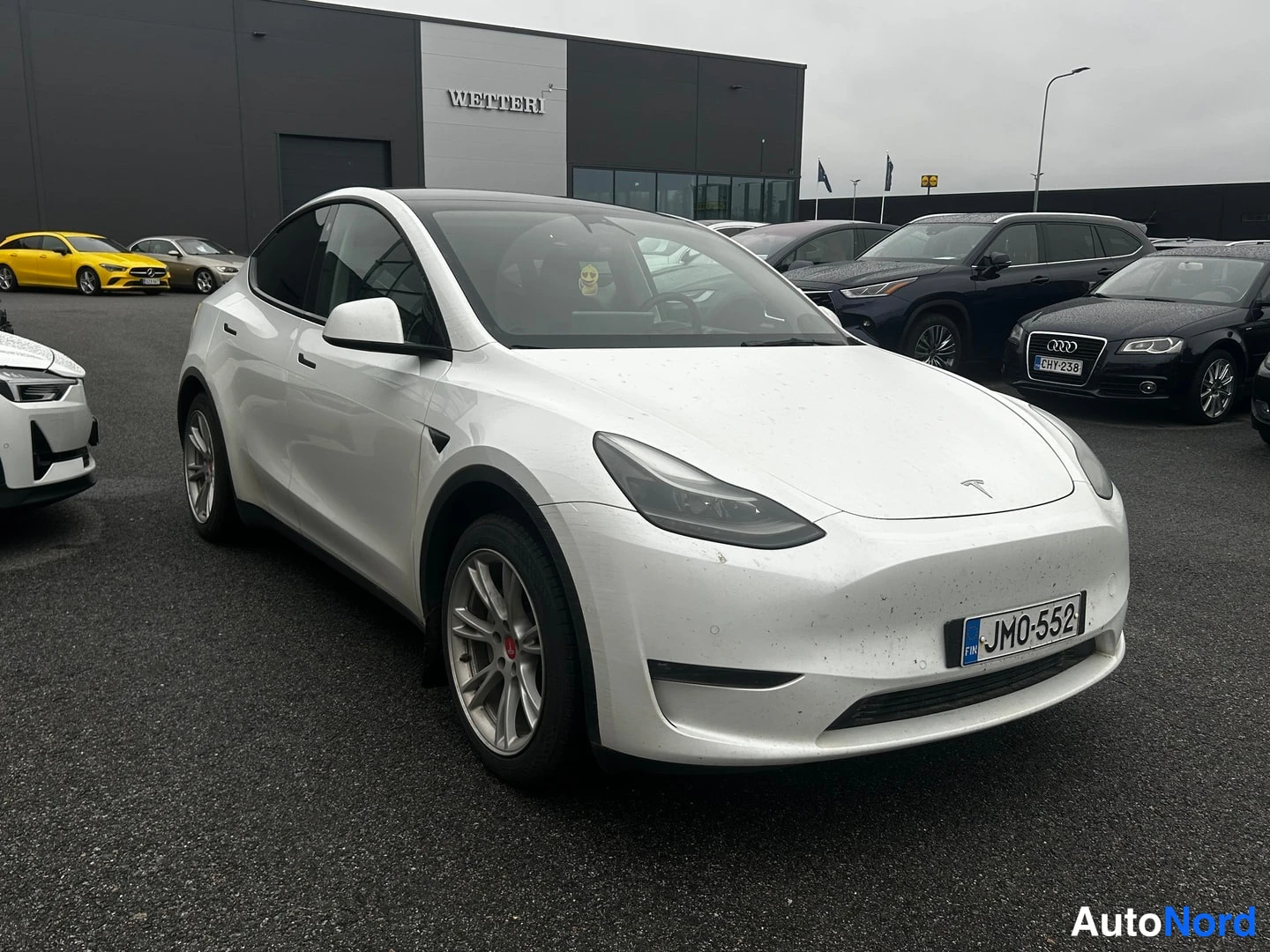 Etuviisto, nokka oikealle: Valkoinen Tesla Model Y etuviistosta vasen kylki näkyvissä, rekisteri, parkkipaikalla.