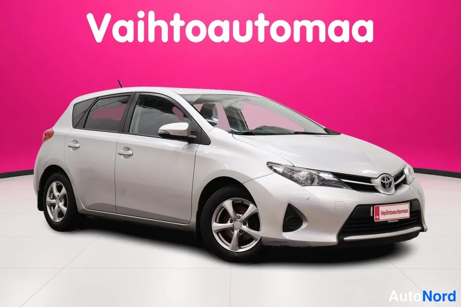 Etuviisto, nokka oikealle: Hopeanvärinen viistosti edestä oikealta kuvattu Toyota Auris -henkilöauto studioavulla vaaleanpunaisella taustalla.