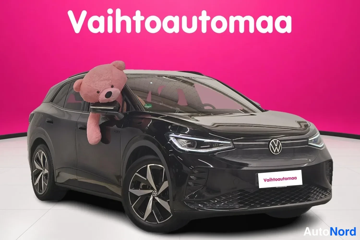 Etuviisto, nokka oikealle: Mustan Volkswagen ID.4:n etunurkkakuva oikealta, keulakilpi ja oikea kylki näkyvissä, suurikokoisen pehmolelunäkin takana katetussa autosalissa.
