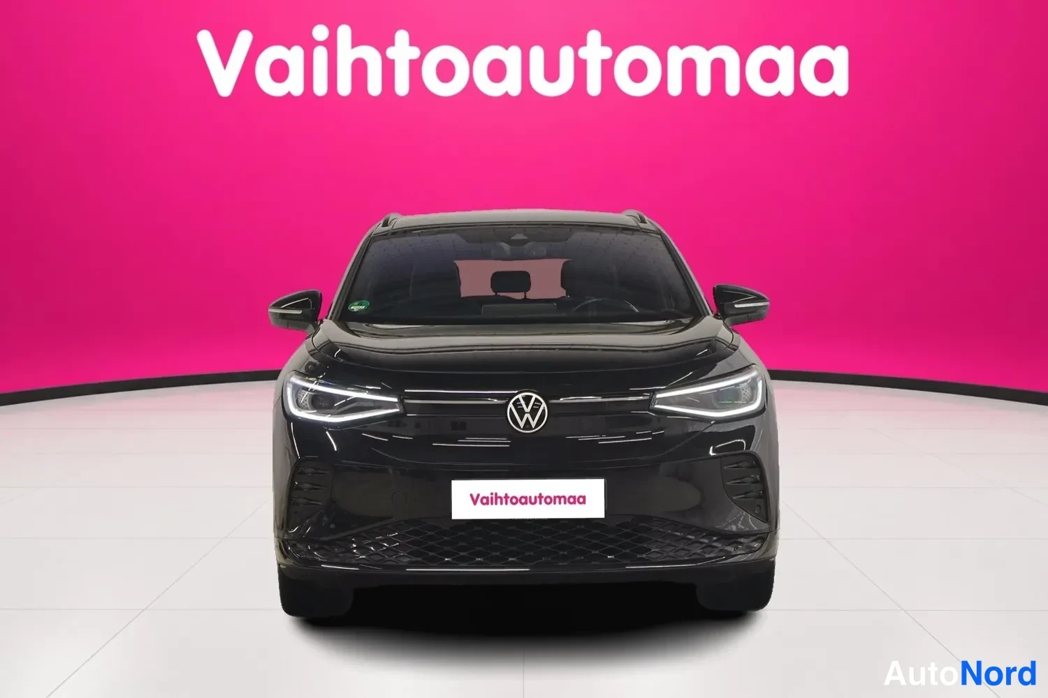 Edestä: Mustan Volkswagen ID.4:n suora etukuva, keula ja etupuskuri keskitettynä katetussa autosalissa.