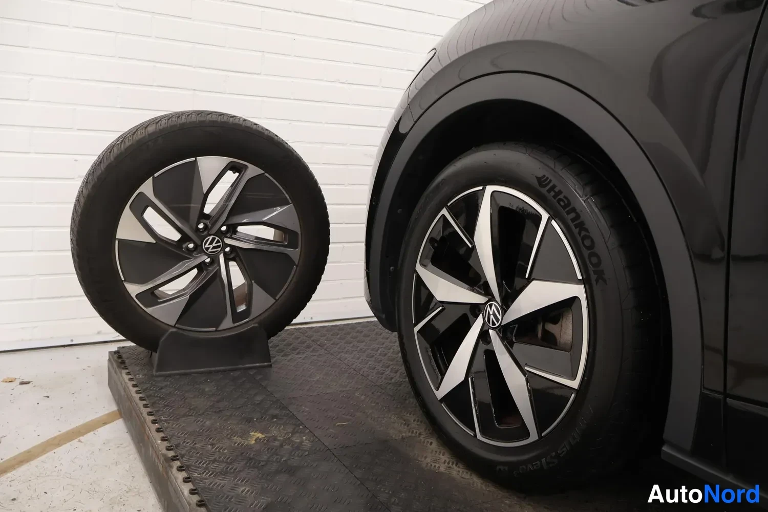 Yksityiskohta: Mustan auton etupyörä valmiilla seoksella vieressä irrallinen varaosa-vanne同样 valmiilla seoksella, molemmissa VW-tunnus ja Hankook-renkaat.