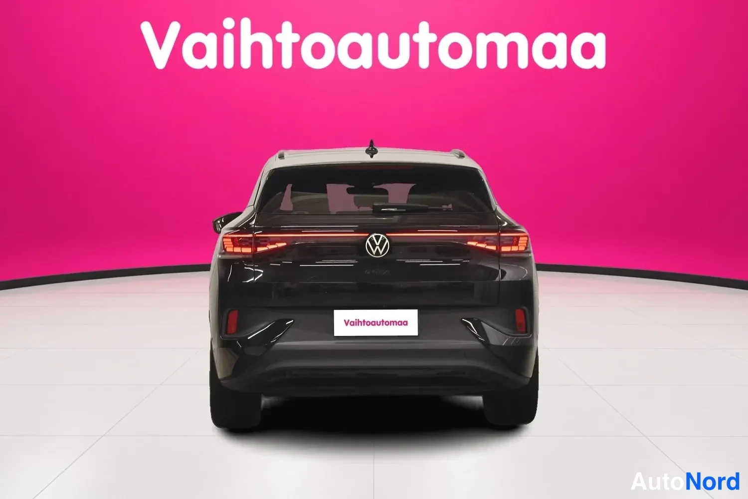 Takaa: Musta VSP suoraan takaa kuvattuna; takavalojen punainen valoviiva, VW‑merkki ja takilasi ovat näkyvissä vaaleanpunaisen taustan edessä.
