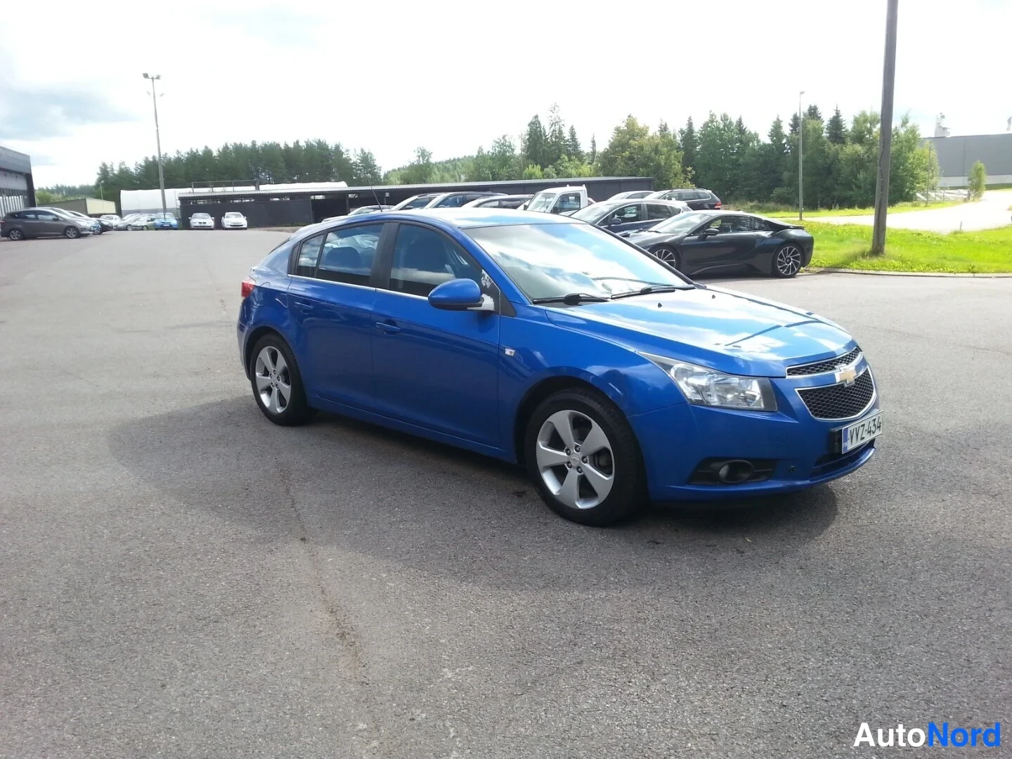 Etuviisto, nokka oikealle: Chevrolet Cruze -viistoperän etukolmasosanäkymä oikeaan nokkaan; auto on sininen ja siinä näkyvät etusivu, etupyörät ja rekisterikilpi.