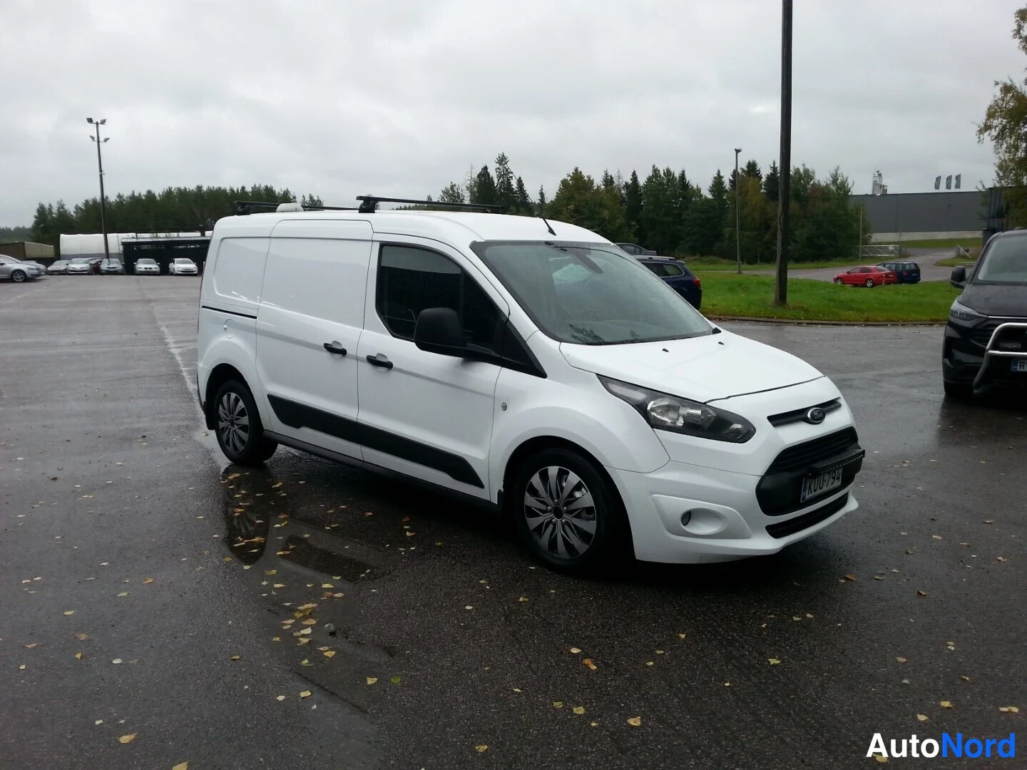Etuviisto, nokka oikealle: Valkoinen Ford Transit Connect -pakettiauto etuosalta ja vasemmalta sivulta, kattoteline asennettuna, märällä parkkipaikalla.