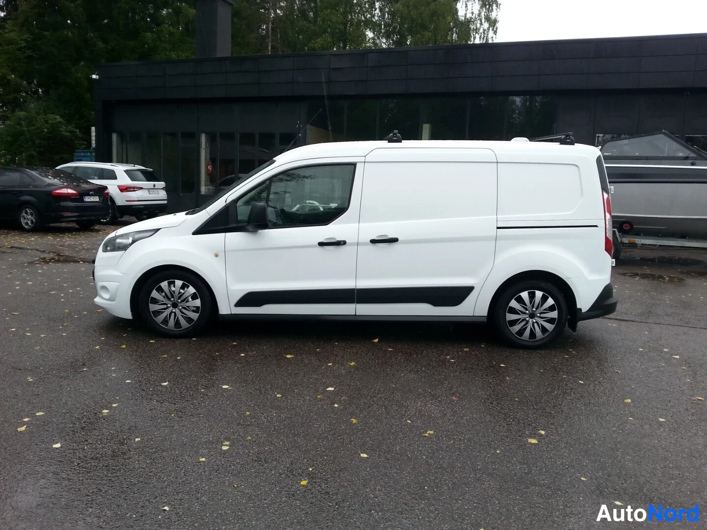 Etuviisto, nokka vasemmalle: Valkoinen Ford Transit Connect -pakettiauto sivulta ja etuosasta, kattoteline asennettuna, pysäköitynä märälle paikalle rakennuksen edessä.