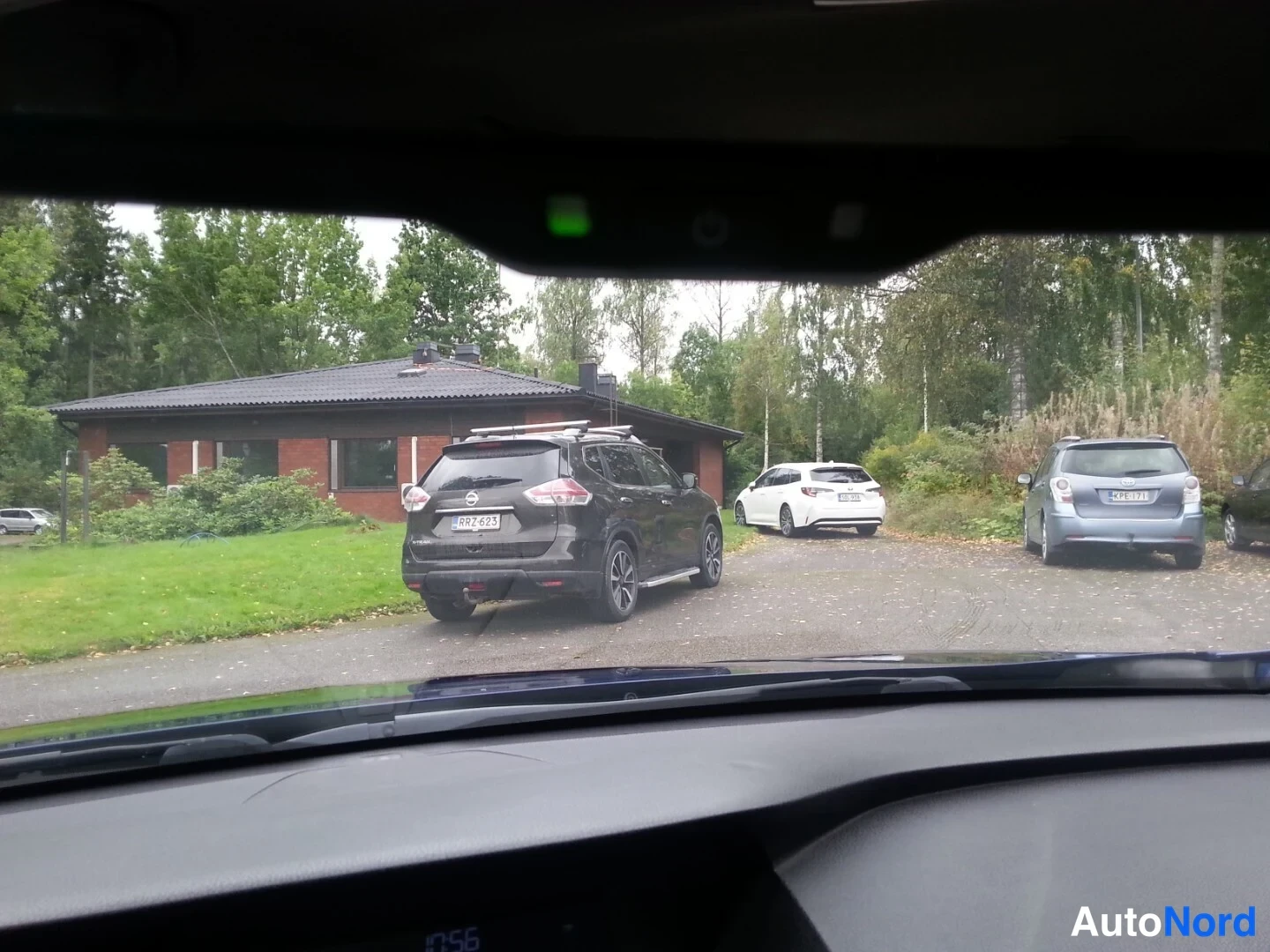 Muu: Näkymä tuulilasin läpi pihaan: punatiilinen talo ja pihalla useita pysäköityjä autoja, edessä musta farmariauto takaa kuvattuna