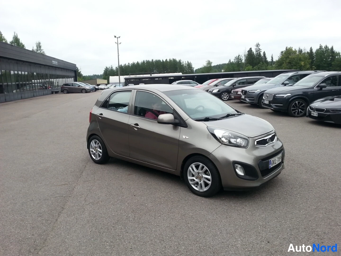 Etuviisto, nokka oikealle: Kia Picanto -henkilöautoa esitetään edestä oikealta kulmasta parkkipihalla, kokonaisuudessaan ruskeanharmaa.