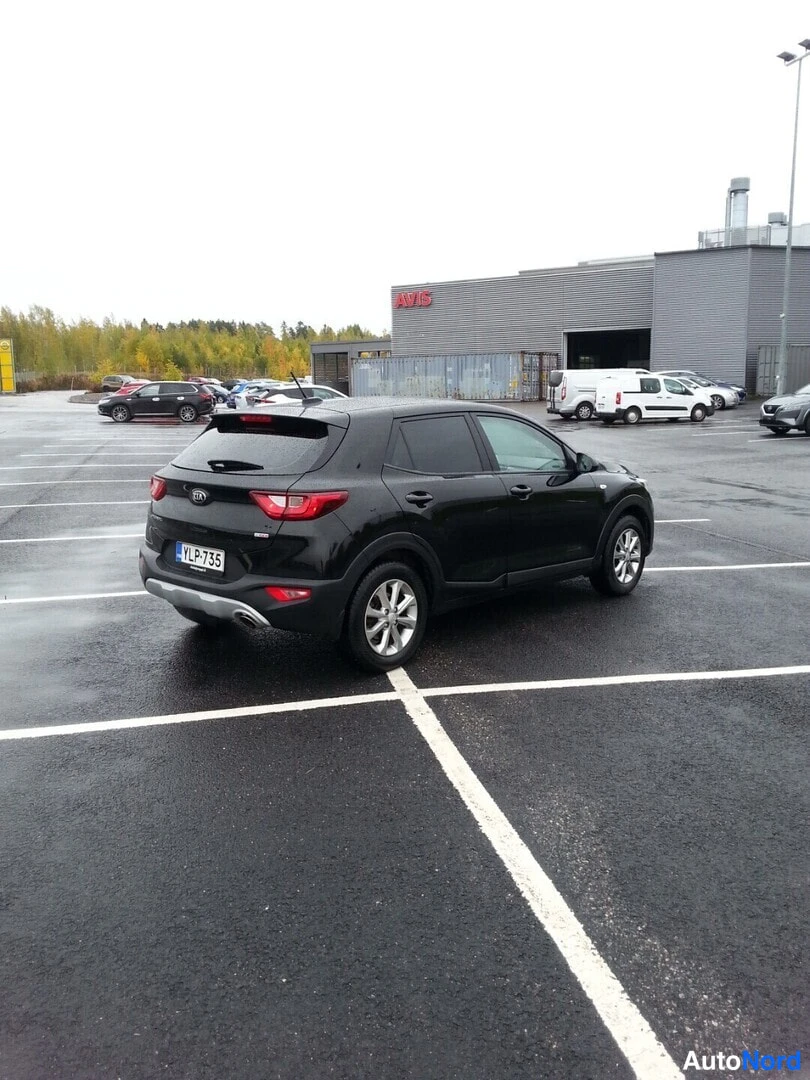 Takaviisto: Kia Stonic -henkilöauto takaa kolmesta neljäsosasta, takapää vasemmalla, mustana parkkipaikalla autotallirakennuksen edustalla.