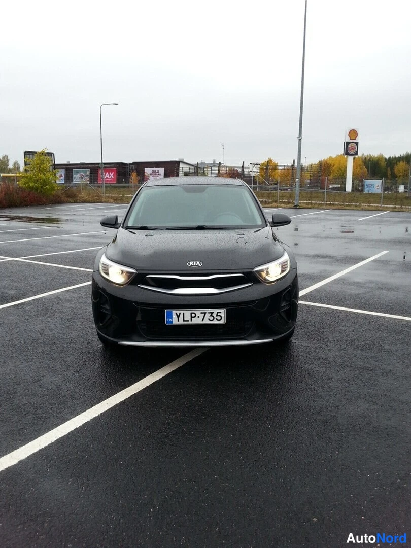 Edestä: Kia Stonic mustana edestäpäin valaistuilla ajovaloilla parkkipaikalla, rekisteritunnus.