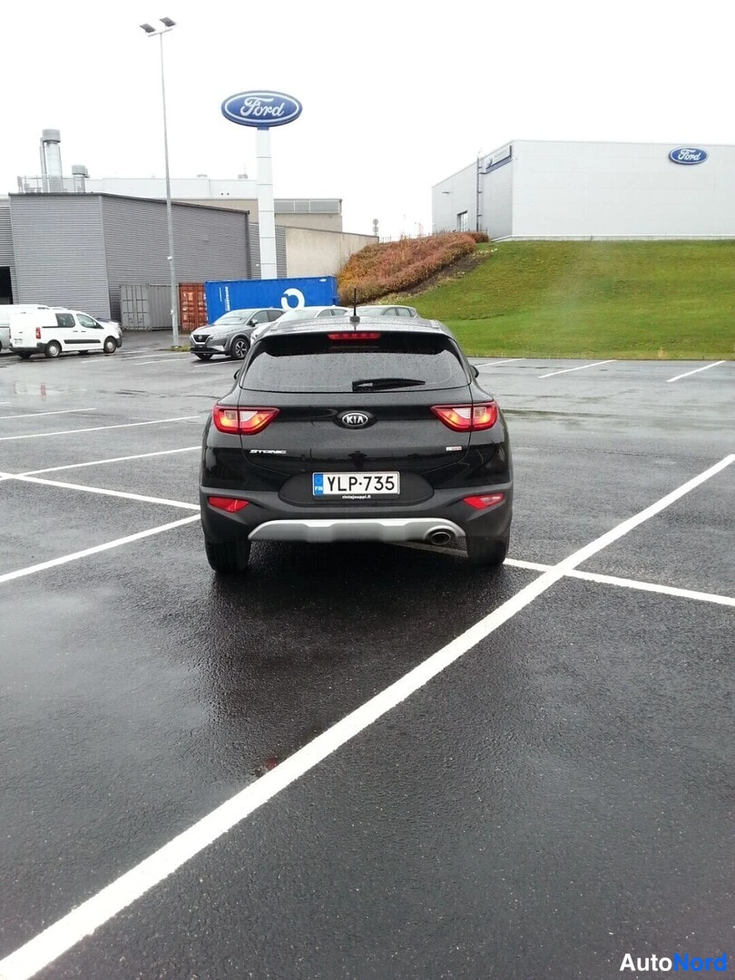 Takaa: Mustan Kia Stonicin takanäkö parkkipaikalla, rekisterikilpi, märkä asfaltti ja Ford-toimipiste taustalla.