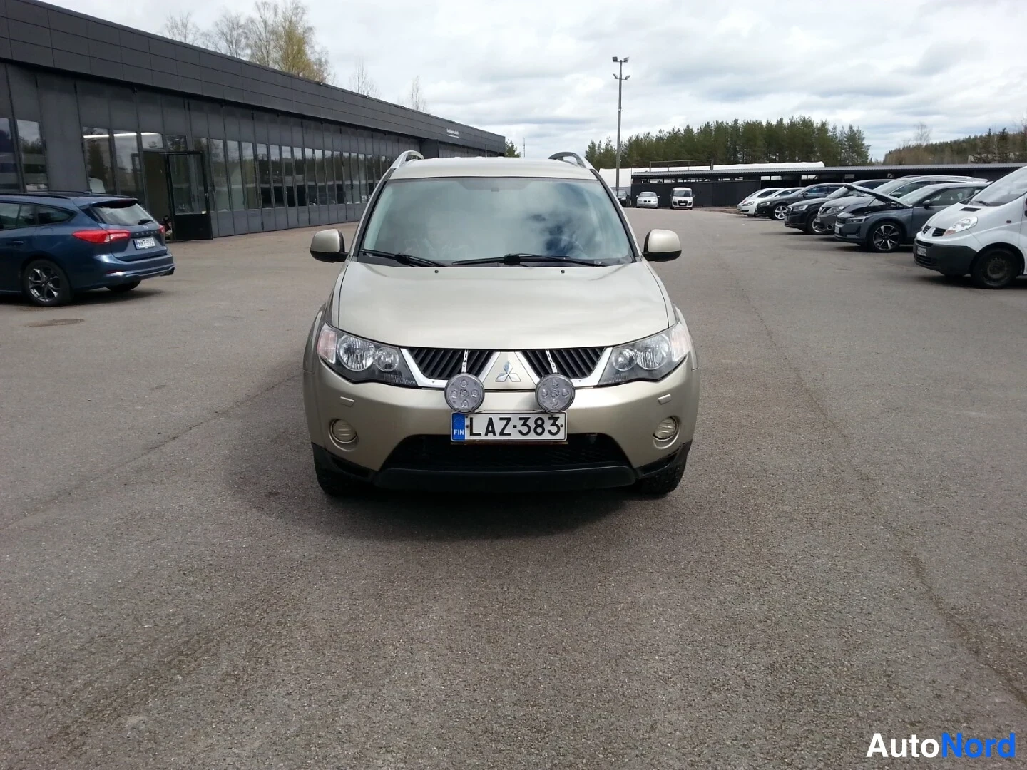Edestä: Beige Mitsubishi Outlander edestäpäin parkkipaikalla, rekisteritunnus, lisävalot etupuskurissa.