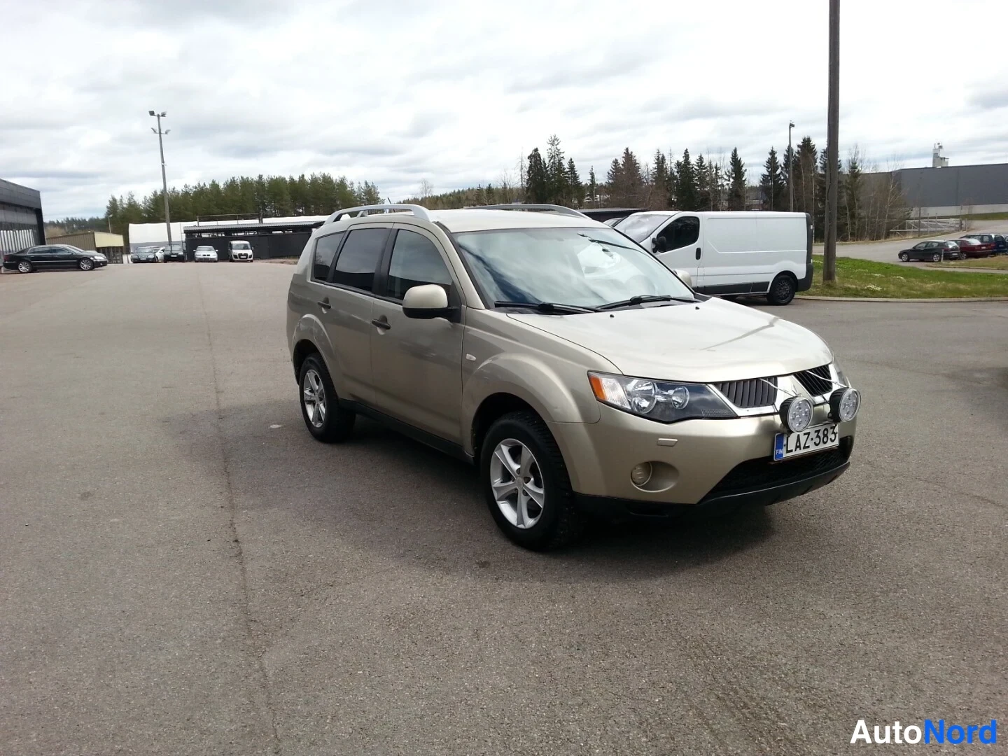 Etuviisto, nokka oikealle: Mitsubishi Outlander -farmarikin etualalla kultainen (beige) maastoauto kolmiväripuolet näkyvät, keula ja etusivu oikealla, rek, lisävalot puskurissa, ka
