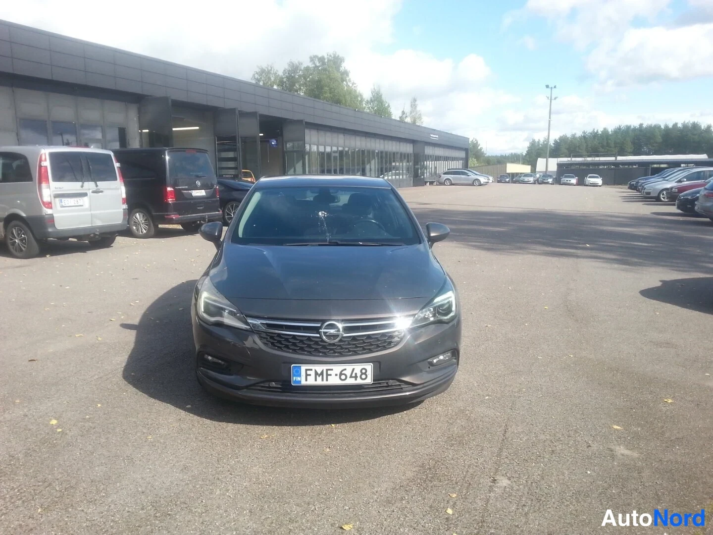 Edestä: Tummanharmaa Opel Astra etukuvalla liiketontin pihalla, rekisteritunnus.
