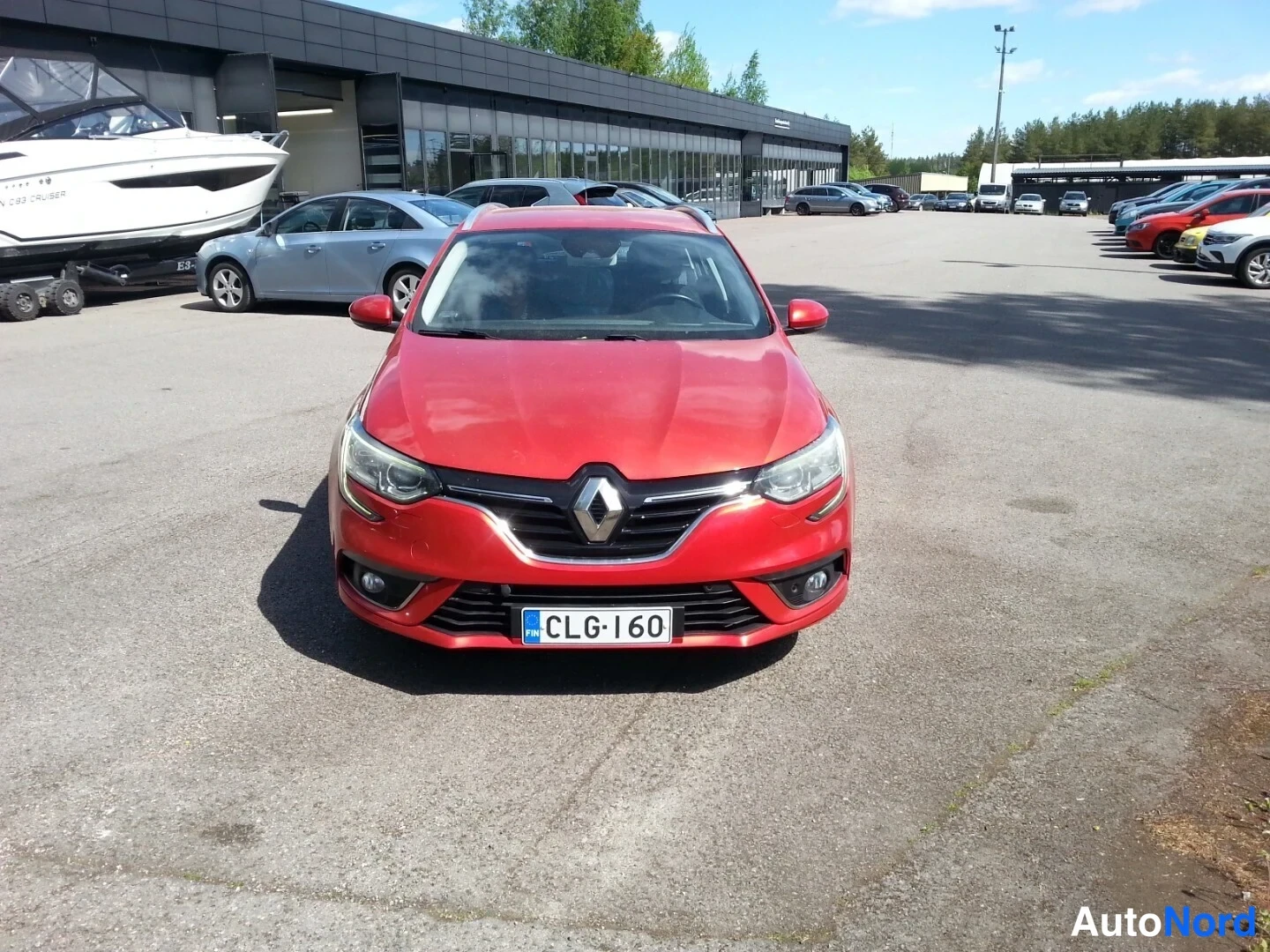 Edestä: punaisen Renault-henkilöauton edestä otettu kuva, auto pysäköitynä huoltamon pihaan, rekisterikilpi, Renault-tunnuksella varustettu etusäleikkö ja sumuvalot